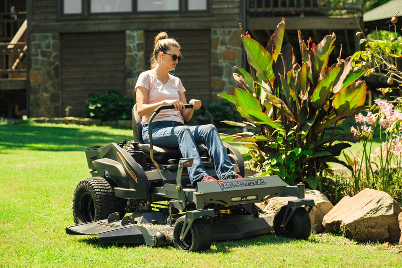 Woman riding spartan mower along flower beds with house in background