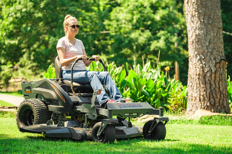 Woman riding spartan mower with flower garden and tree in background